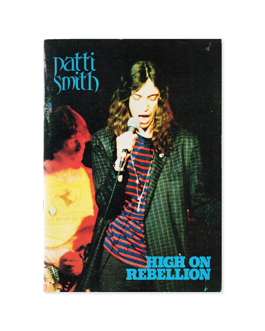 A vintage Patti Smith 'High on Rebellion' booklet photographed flat against a white background. The cover features a dark concert scene with Patti Smith singing into a microphone, a guitarist partially visible on the left, and bright blue text reading 'patti smith' and 'HIGH ON REBELLION'.