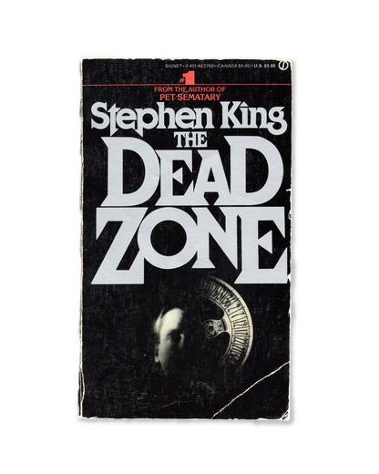 The cover of the paperback book 'The Dead Zone' by Stephen King, featuring a black background with the title in white letters and a image of a skull in the center.